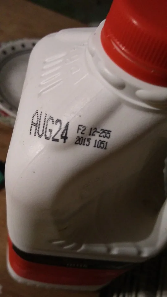 A close-up of a white plastic bottle with a red cap, showing a printed date 'AUG24' and other numbers 'F2 12-255 2015 1051'…
