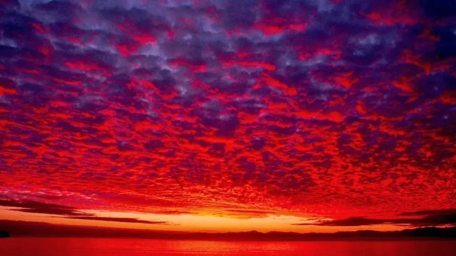 A dramatic sky filled with fiery red and deep purple clouds over a calm body of water at sunset or sunrise. The horizon…