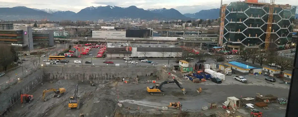 An aerial photo of a large urban construction site with excavators and a deep excavation pit, surrounded by roads, parked…