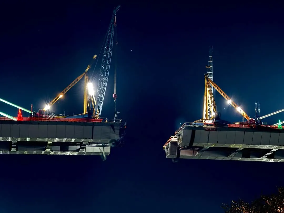Two massive bridge segments hang suspended in the night sky, each illuminated by construction lights and cranes, poised to…