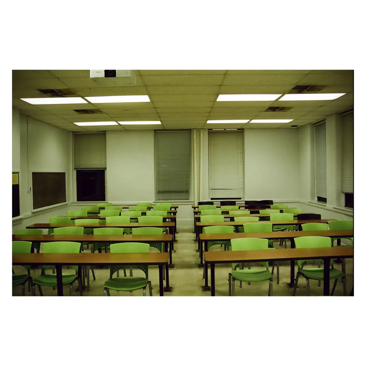An empty classroom with rows of green chairs and wooden desks under fluorescent lights, with a blackboard on the left wall…