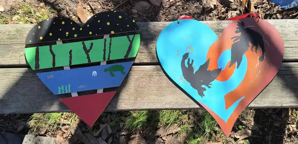 Two hand-painted wooden hearts sit side-by-side on a park bench. The left heart is segmented into layers showing a night sky…