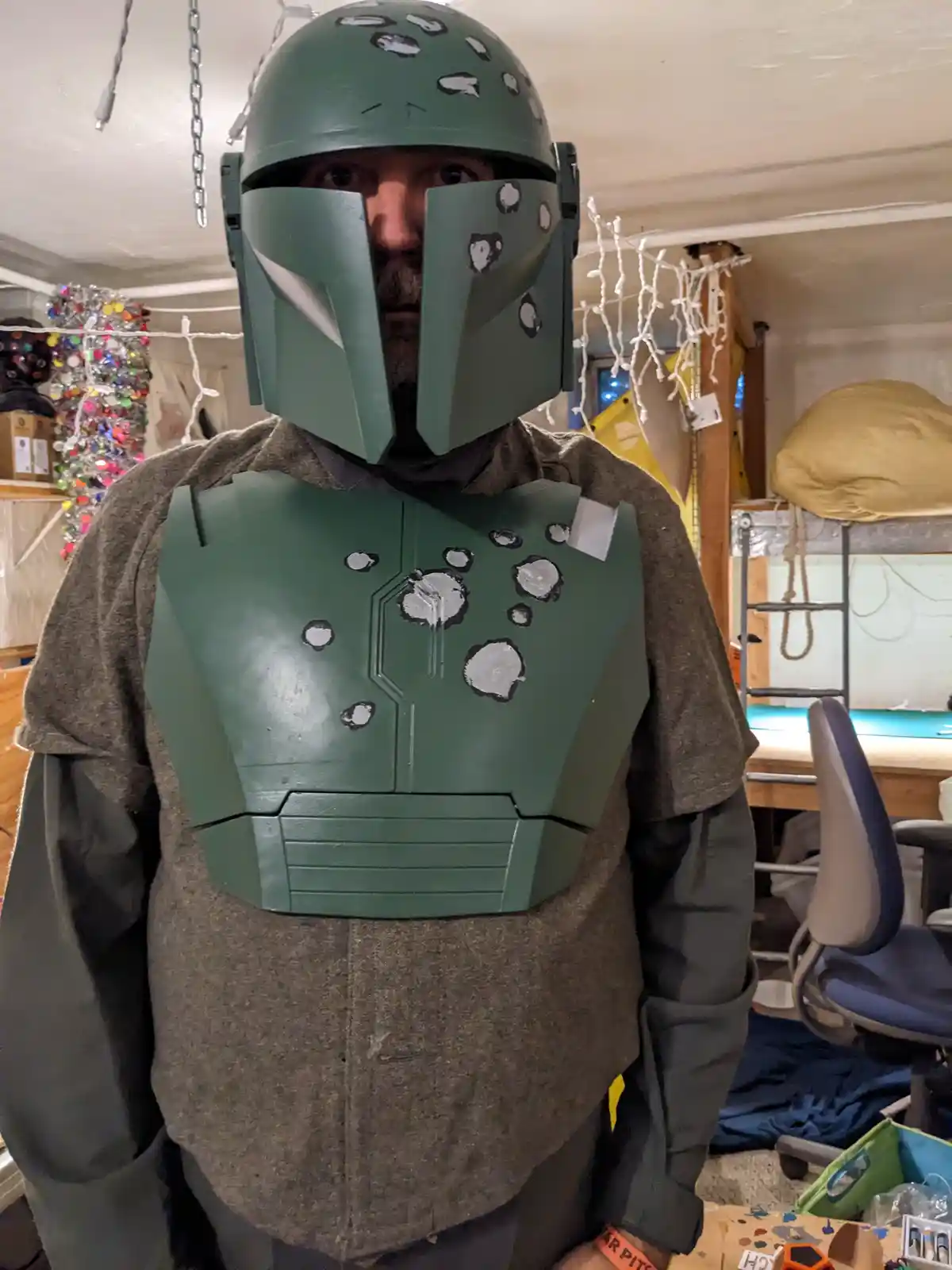 A man in a green Boba Fett costume with numerous white painted bullet holes on the helmet and chest plate stands in a…
