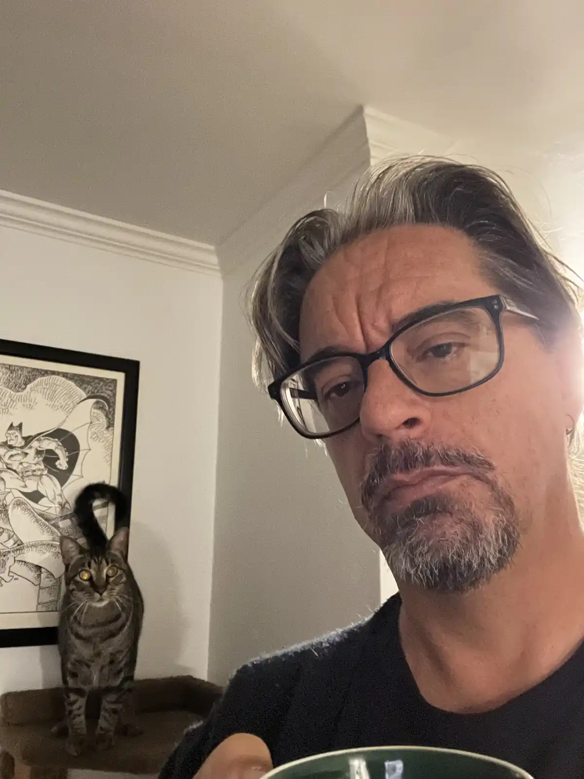 A man with glasses and a graying beard stares intently at the camera while holding a green mug, his expression conveying…