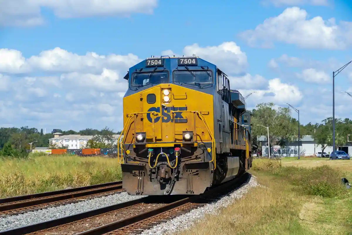 A blue and yellow CSX freight train engine, numbered 7504, is shown on railway tracks under a partly cloudy sky. The train…