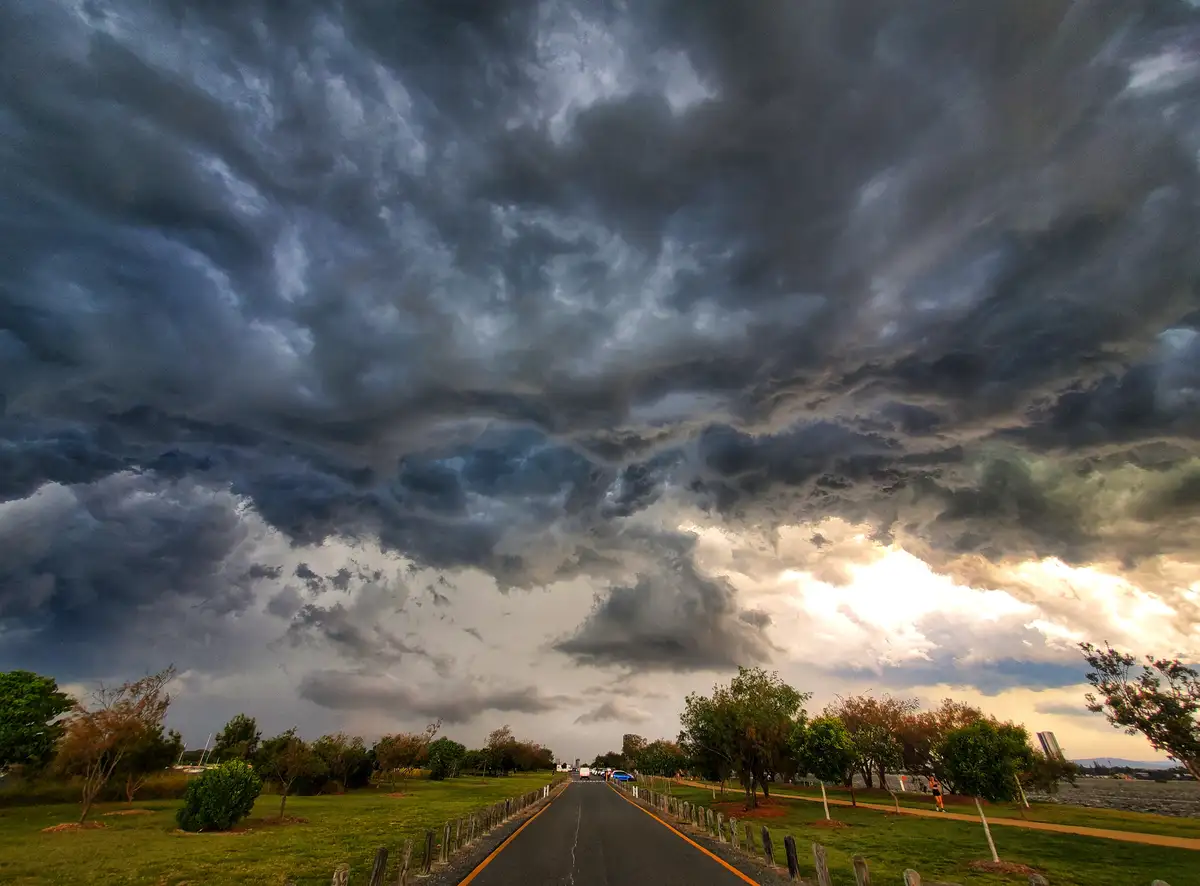 A dramatic landscape photo shows a long road stretching toward a horizon under a massive, dark, stormy sky with ominous…