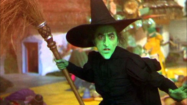 A green-skinned witch in a black pointed hat and robe holds a broomstick, looking shocked or surprised. The overlaid text…