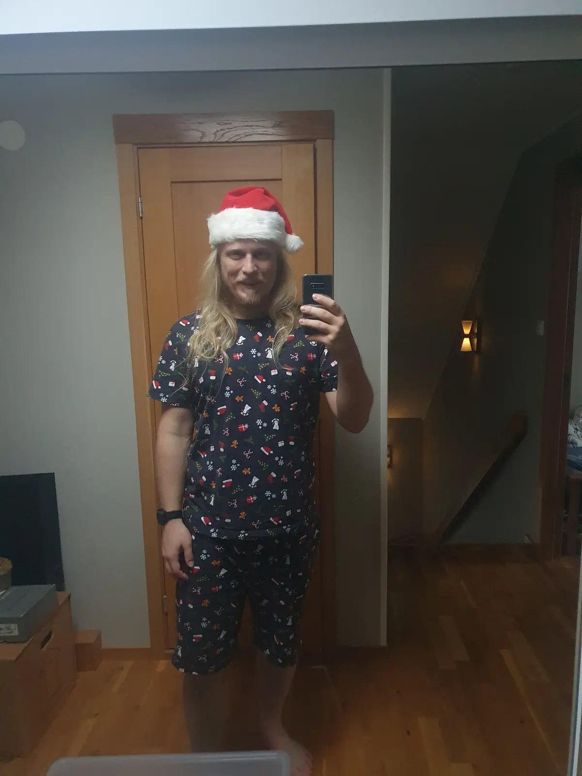 A man with long blonde hair and a beard takes a mirror selfie while wearing a red and white Santa hat and matching dark blue…