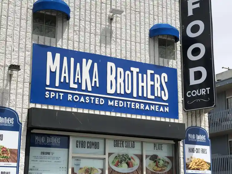 A photo of the exterior of a restaurant named 'Malaka Brothers Spit Roasted Mediterranean' with a blue sign and menu items…