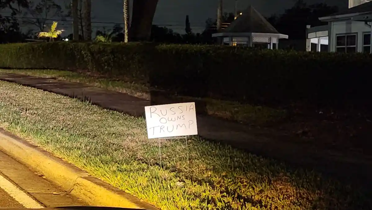 A hand-written sign on a lawn at night reads 'RUSSIA OWNS TRUMP'. The sign is placed in a grassy area beside a sidewalk…