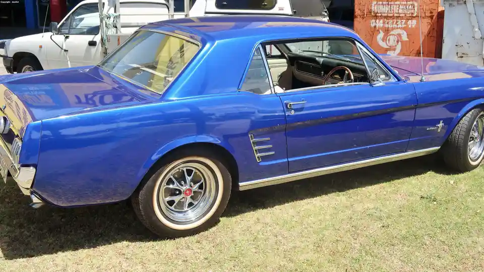 A shiny blue classic Ford Mustang coupe is parked on grass, its chrome details and whitewall tires gleaming under sunlight…