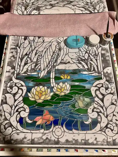 A stained glass art project in progress, showing a pond scene with lily pads, water lilies, and a fish, where most sections…
