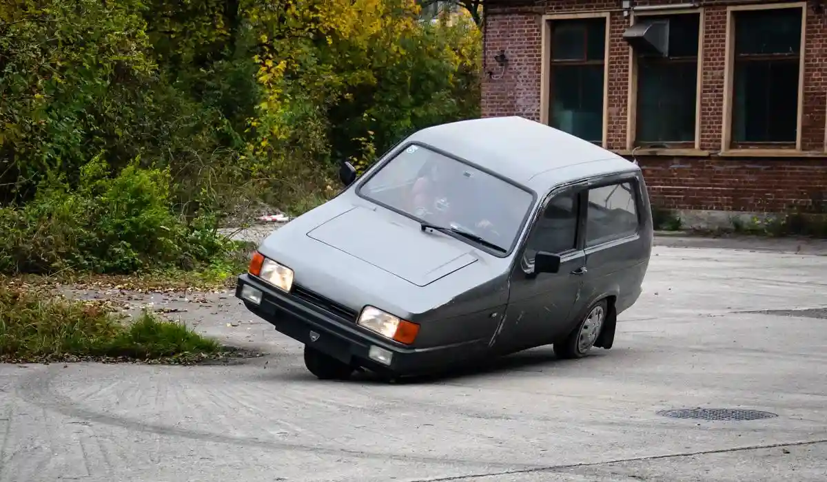a gray, boxy, vintage van is parked at an odd angle on a concrete surface, appearing to lean precariously to one side. The…