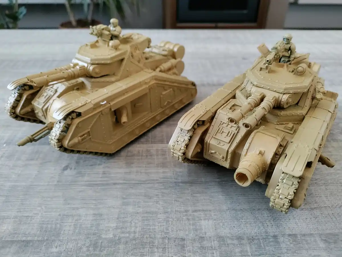 Two unpainted, tan-colored plastic model tanks sit side-by-side on a gray textured table, each with a small crew figure on…