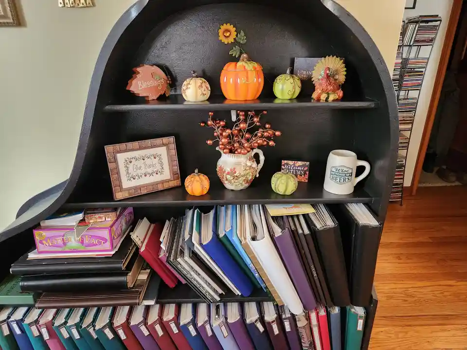 A black arched bookshelf is decorated with autumn-themed items including pumpkins, a turkey figurine, and signs reading…