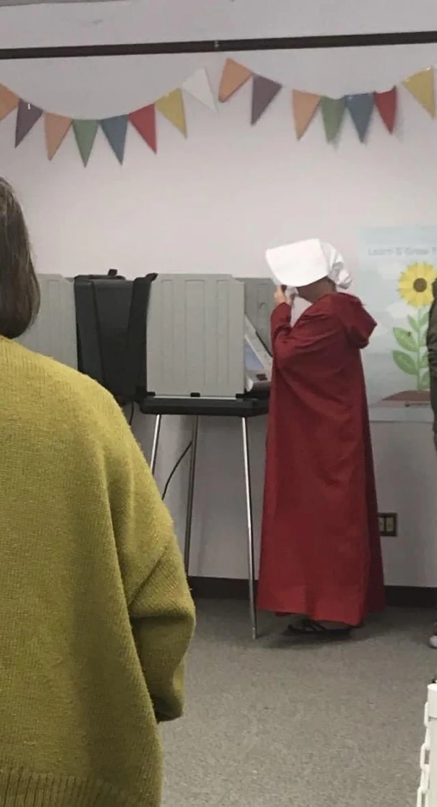 A person dressed in a red robe and white bonnet, resembling a Handmaid from 'The Handmaid's Tale,' stands at a voting booth…
