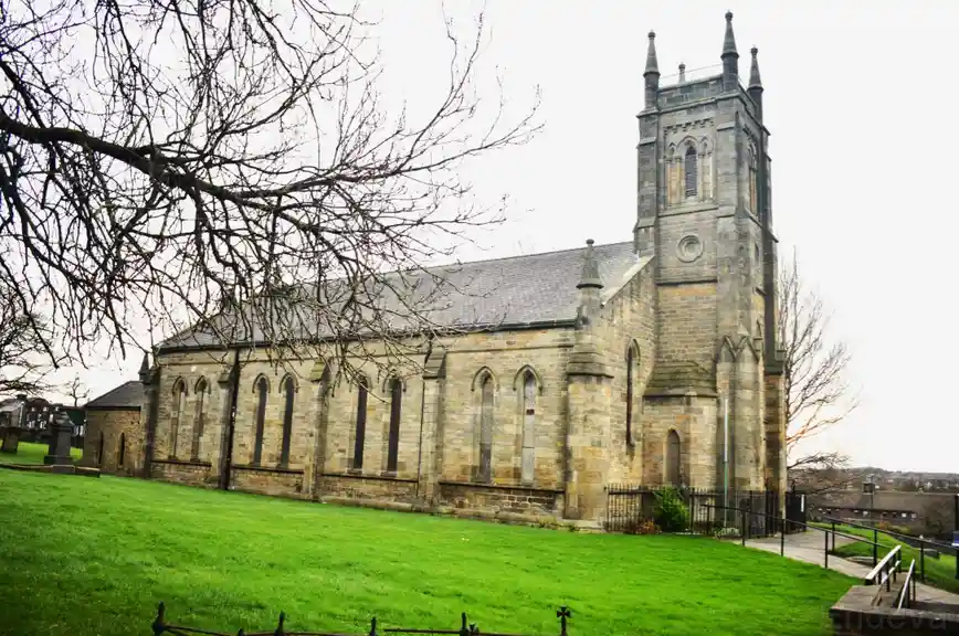 A large stone church with a tall tower and arched windows stands on a grassy hill, framed by bare tree branches against an…