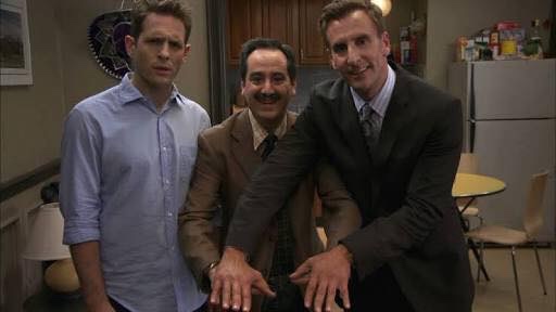 Three men stand together in a kitchen-like setting. The man on the left looks uncomfortable and annoyed, the man in the…