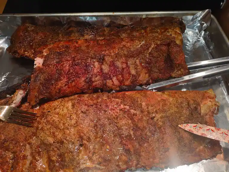Three large racks of seasoned, cooked ribs rest on aluminum foil in a metal baking tray. A metal fork is stuck into the…