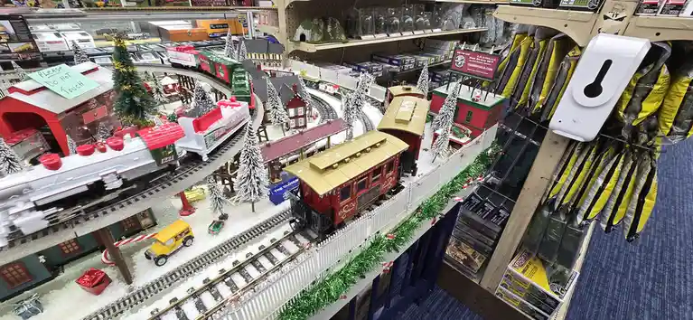 A detailed model train display in a hobby shop, featuring multiple tracks with Christmas-themed trains, snow-covered…