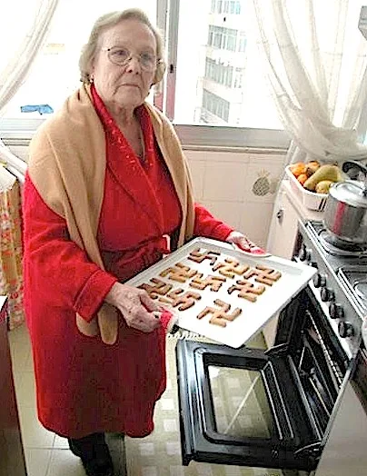 An elderly woman wearing a red robe and beige shawl stands in a kitchen holding a baking tray filled with cookies shaped…
