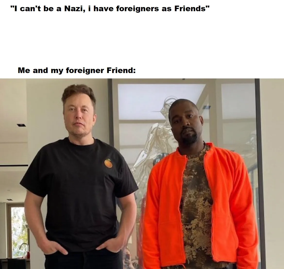 Two men stand side by side indoors; the man on the left wears a black t-shirt with a small orange logo and has his hands in…
