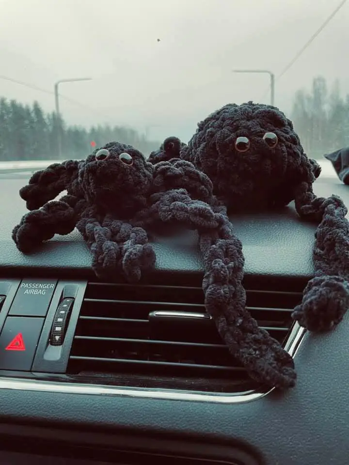 Two black, crocheted spider plushies with large googly eyes sit on a car dashboard, one partially draped over the air vent…