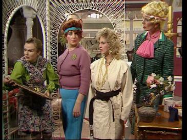 Four women in colorful, 1970s-style costumes stand in a room with ornate mirrors and furniture. The woman on the far left…