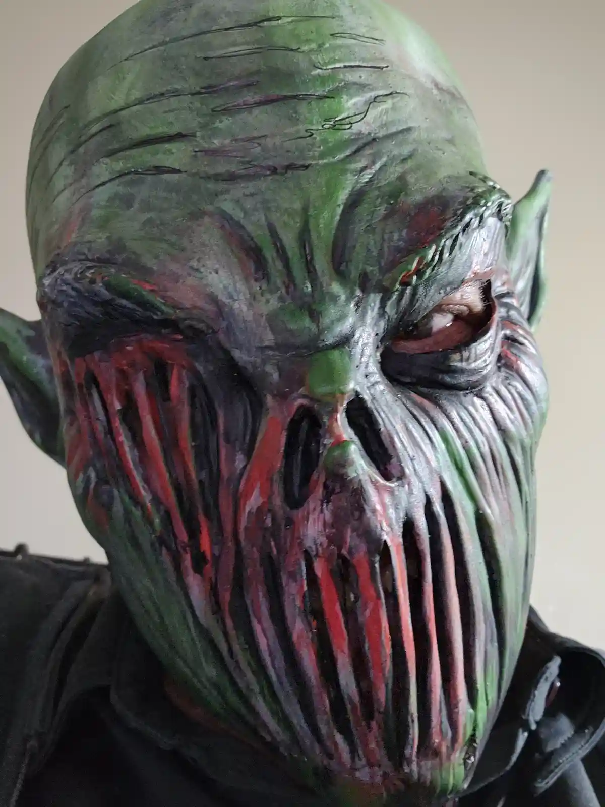 A close-up of a green-skinned, goblin-like creature with pointed ears, deep wrinkles, and a face painted with red streaks…