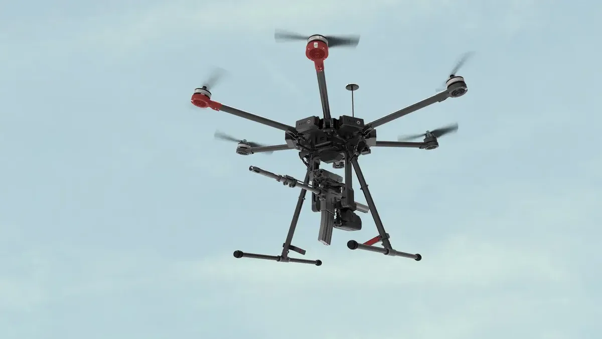 A black hexacopter drone with red motor housings hovers against a pale blue sky, visibly equipped with a mounted machine gun…