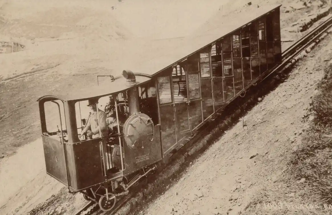 A sepia-toned historical photograph shows an old steam-powered incline railway car ascending a steep, narrow track. Two men…