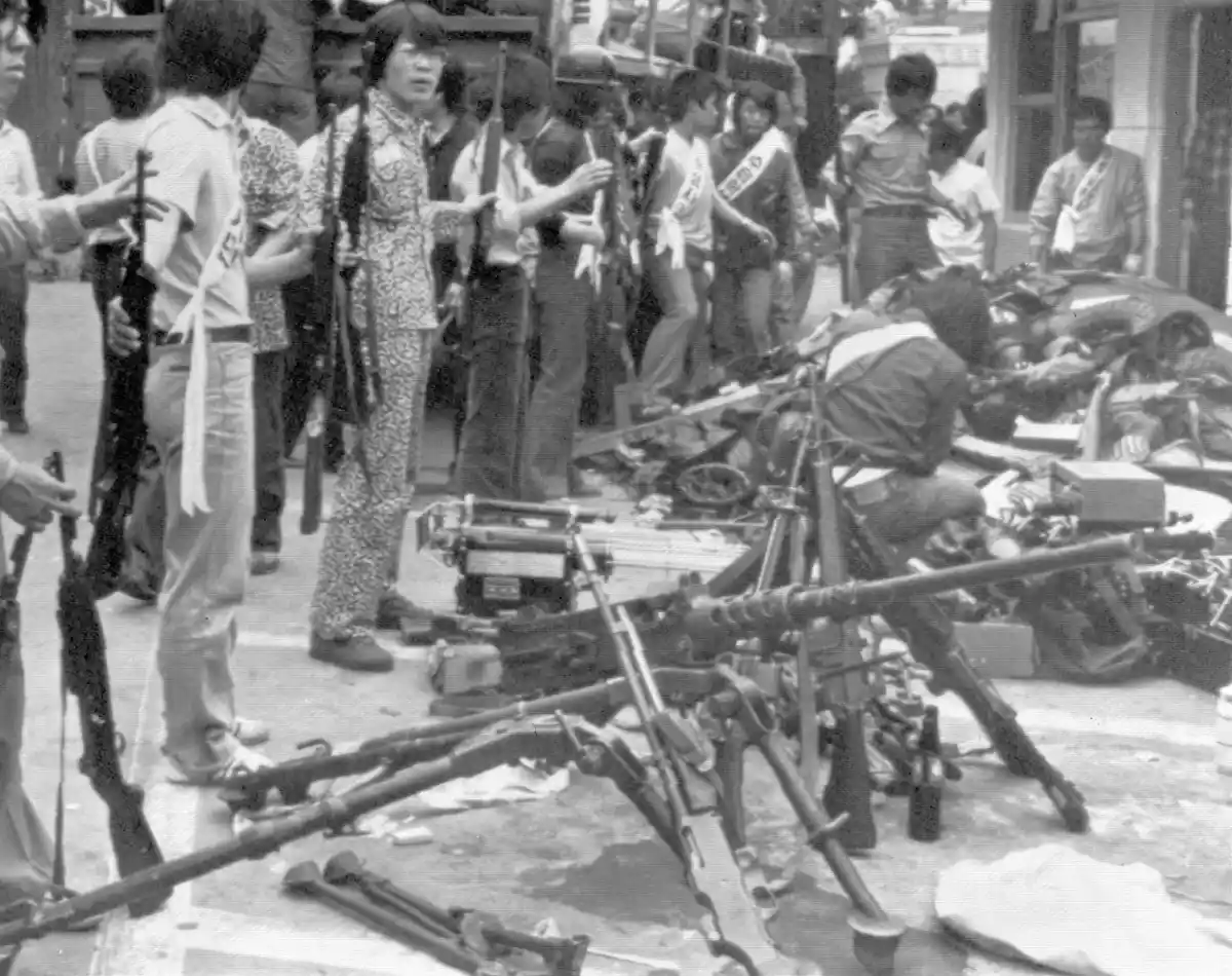 A black-and-white historical photograph shows a group of young people, some wearing sashes, standing among numerous rifles…