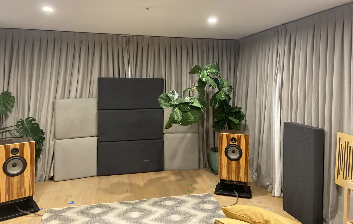 A room with light wood flooring and grey curtains, featuring two large wooden speakers flanking a central arrangement of…