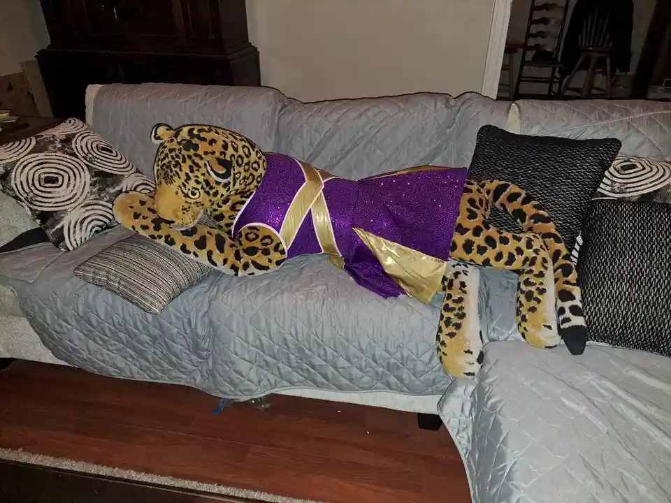 A large plush leopard toy wearing a purple and gold cheerleader outfit is lying sprawled across a gray quilted couch…