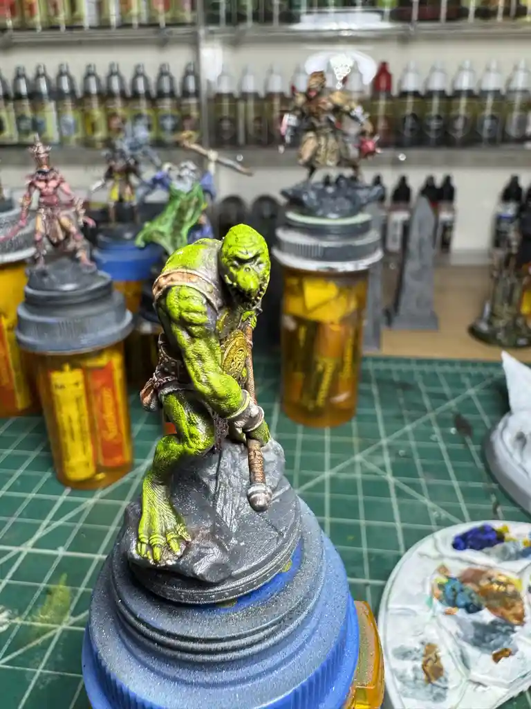 A close-up photo shows a green-skinned, muscular fantasy miniature figure with a fierce expression, standing on a grey base…