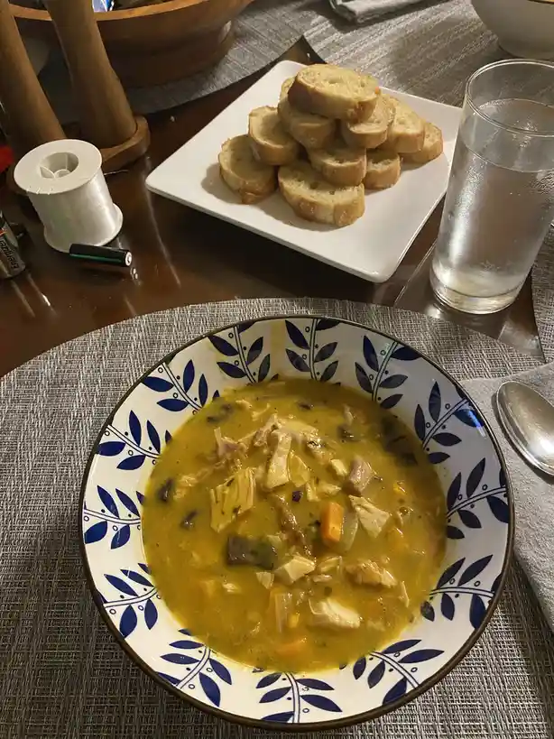 A close-up photo shows a bowl of yellowish chicken and vegetable soup with chunks of meat and carrots, served in a white…