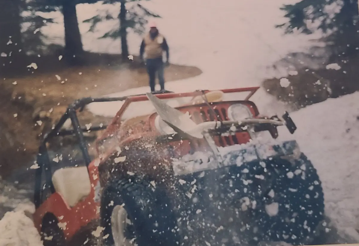 A blurry, snowy photo shows a red off-road vehicle, possibly a Jeep, kicking up snow as it drives through a snowy forest…