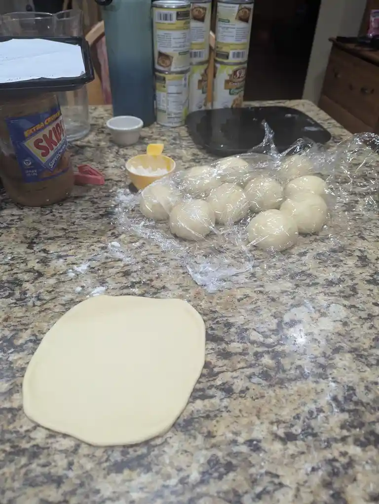 A kitchen counter with a flattened piece of dough in the foreground and a plastic-wrapped batch of dough balls behind it. A…