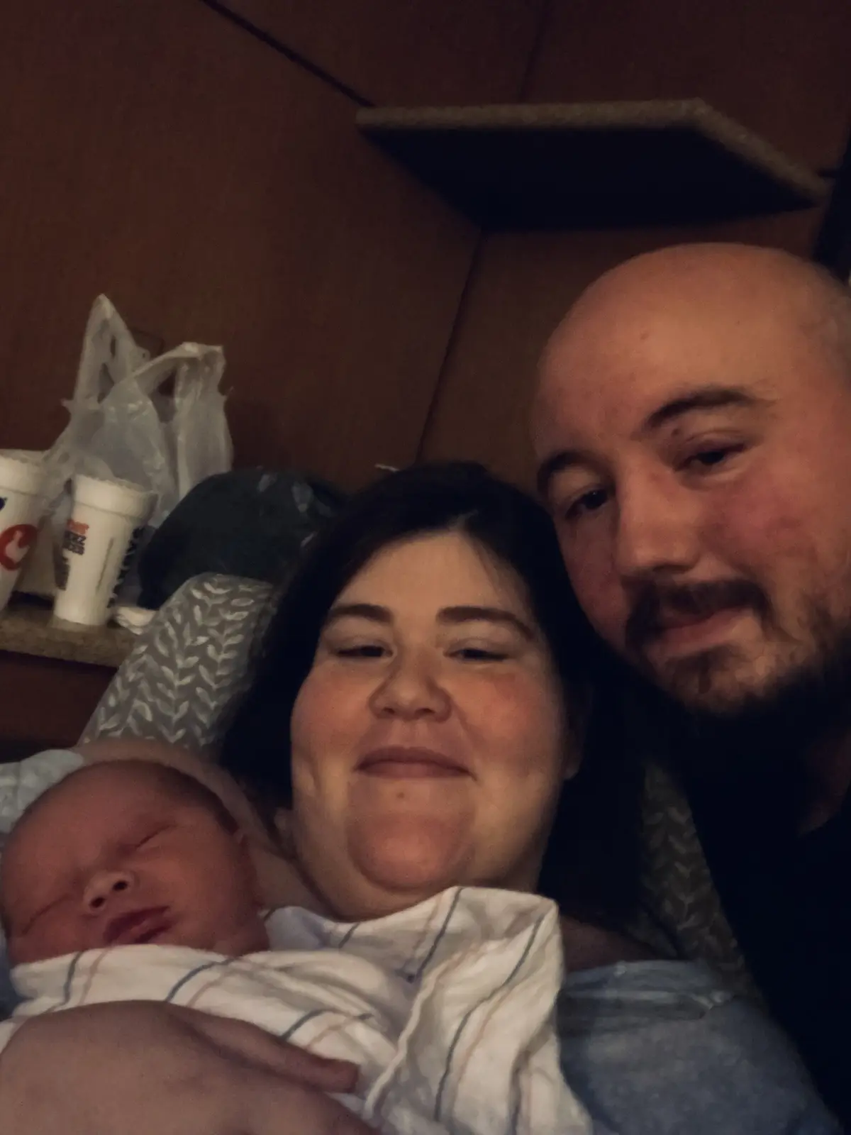 A tired-looking woman with dark hair holds a sleeping newborn wrapped in a striped blanket, while a bald man with a beard…