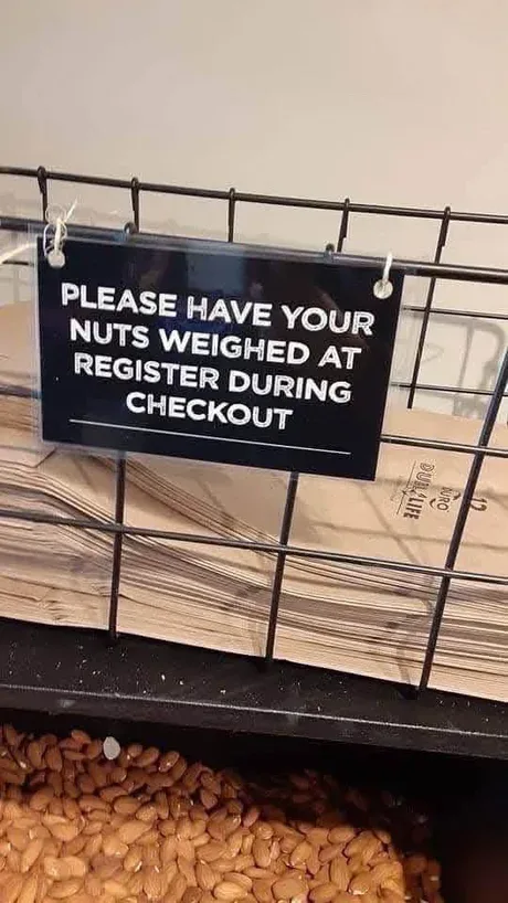 A black sign with white text hangs on a wire rack above a bin of almonds, instructing customers to have their nuts weighed…
