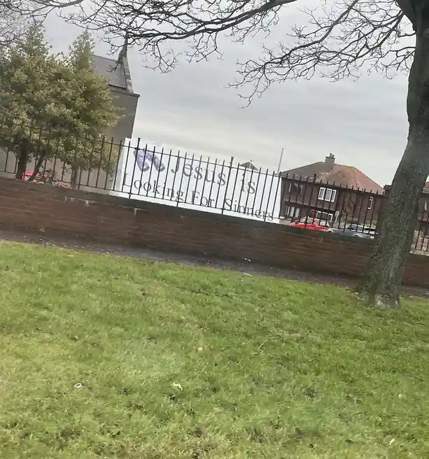 A white banner with the text 'Jesus is Looking For Sinners' is attached to a black metal fence atop a red brick wall. The…