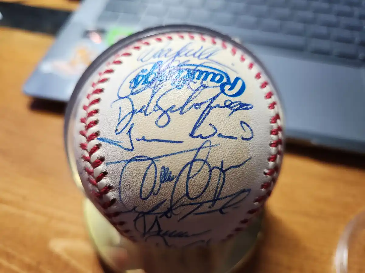 A close-up photo shows a baseball covered in numerous blue ink signatures, resting on a wooden surface with a laptop…