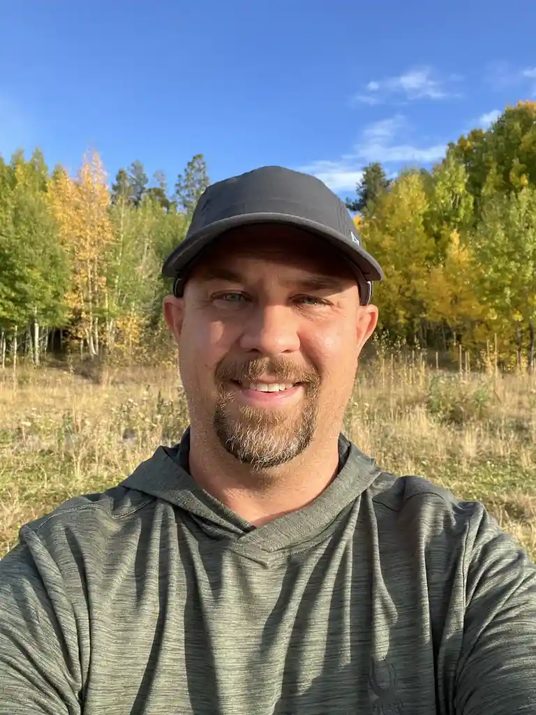 A smiling man with a beard and a black cap takes a selfie outdoors in front of autumn-colored trees under a clear blue sky…