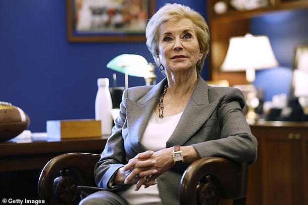 a woman with short blonde hair, wearing a grey blazer over a white top, sits in a wooden chair with her hands clasped…