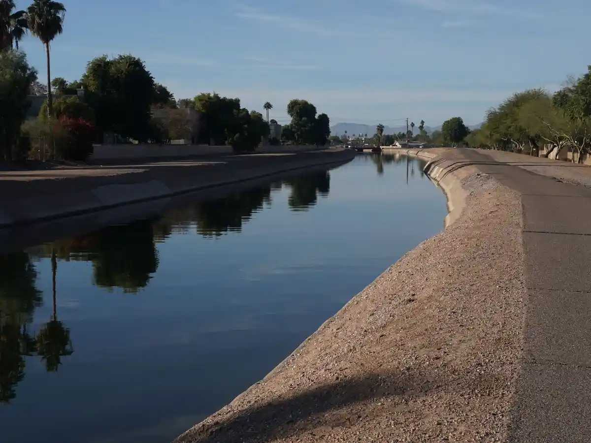 A calm, straight canal with concrete banks and gravel paths under a clear blue sky, flanked by trees and distant mountains…