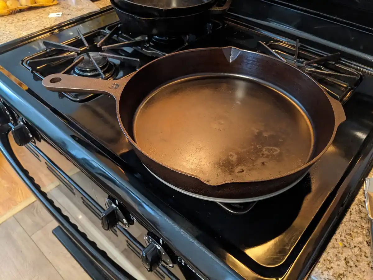 A well-seasoned cast iron skillet sits on a black gas stove burner, its surface showing signs of use with a slightly uneven…