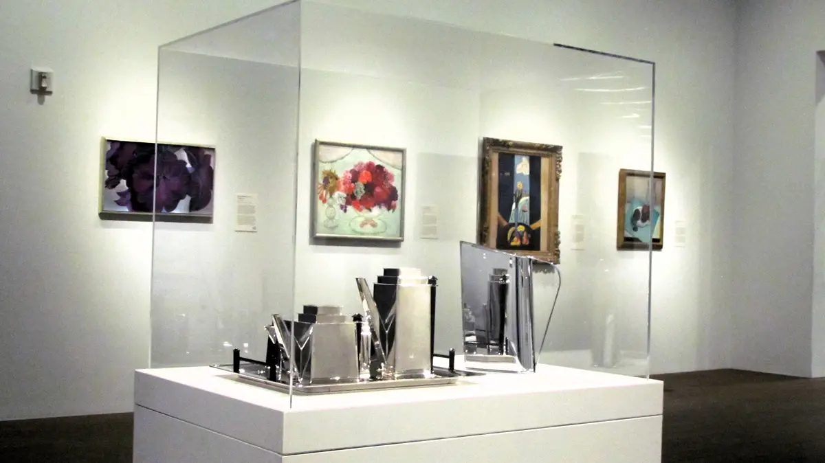 A museum gallery with several framed paintings on the walls behind a glass barrier. In the foreground, a white pedestal…