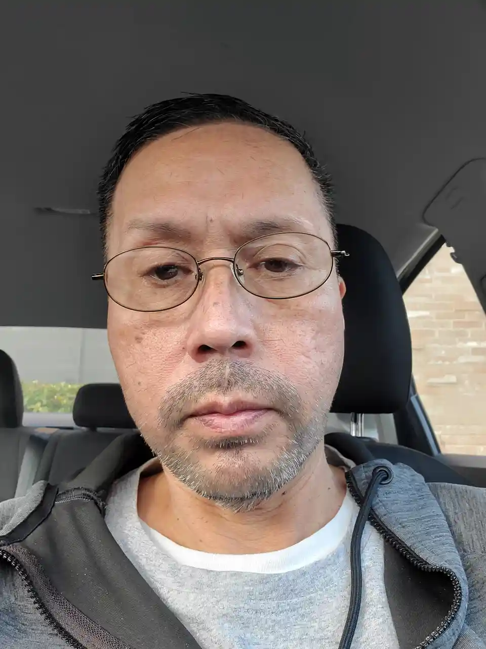A man with glasses and a gray hoodie sits in a car, looking directly at the camera with a neutral expression. The overlaid…
