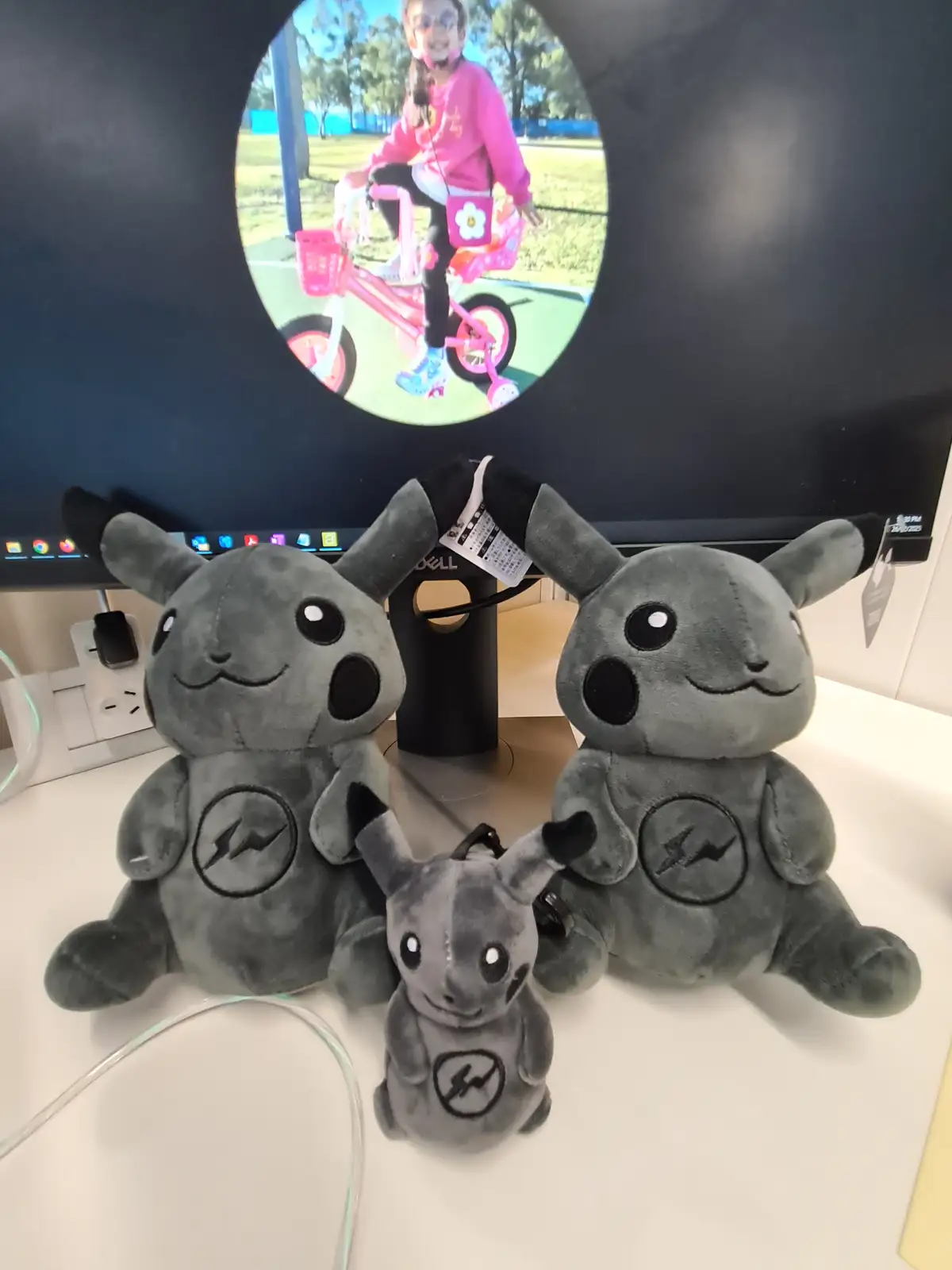 Three grey plush Pikachu toys sit on a desk in front of a monitor displaying a photo of a young girl on a pink tricycle. The…