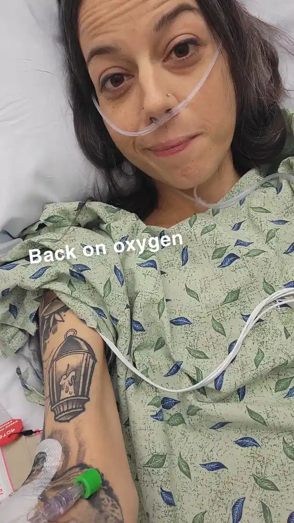 A woman in a hospital gown with a nasal cannula and IV line looks at the camera with a weary but amused expression. Her left…